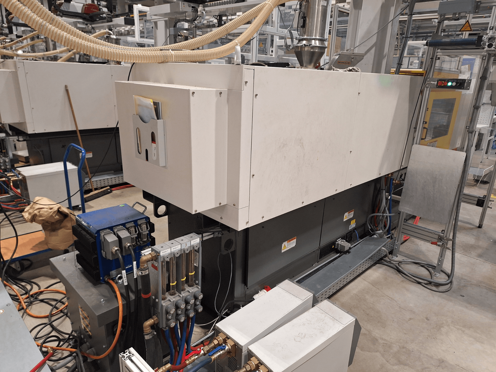 Equipo industrial en una fábrica; incluye una gran máquina con panel de control, mangueras y conexiones eléctricas, vista lateral.