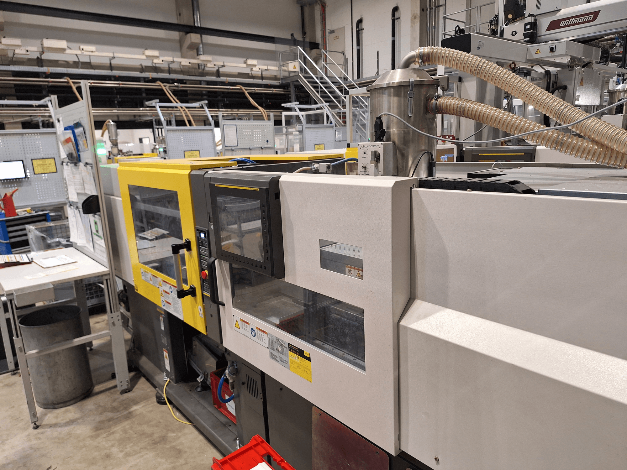 Vista frontal de una máquina de moldeo por inyección multicolor, con secciones amarillas y grises, con panel de control y sistema de escape.